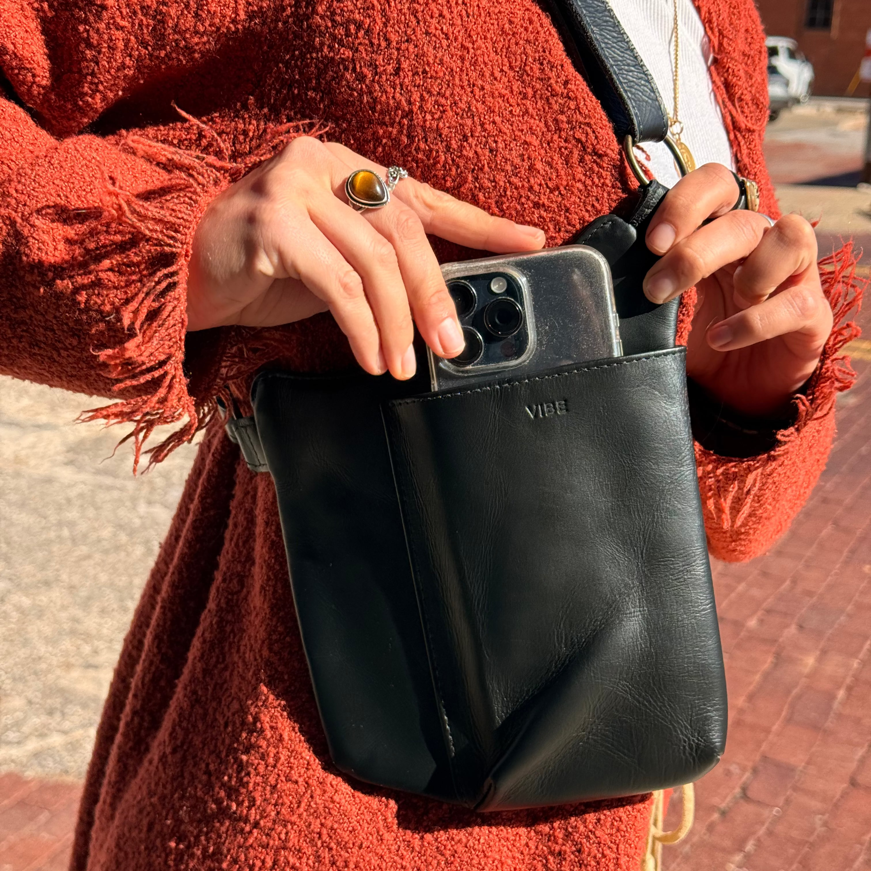 Person holding a black leather bag with a visible brand logo, wearing a red coat. VIBE lifestyle leather bag 
