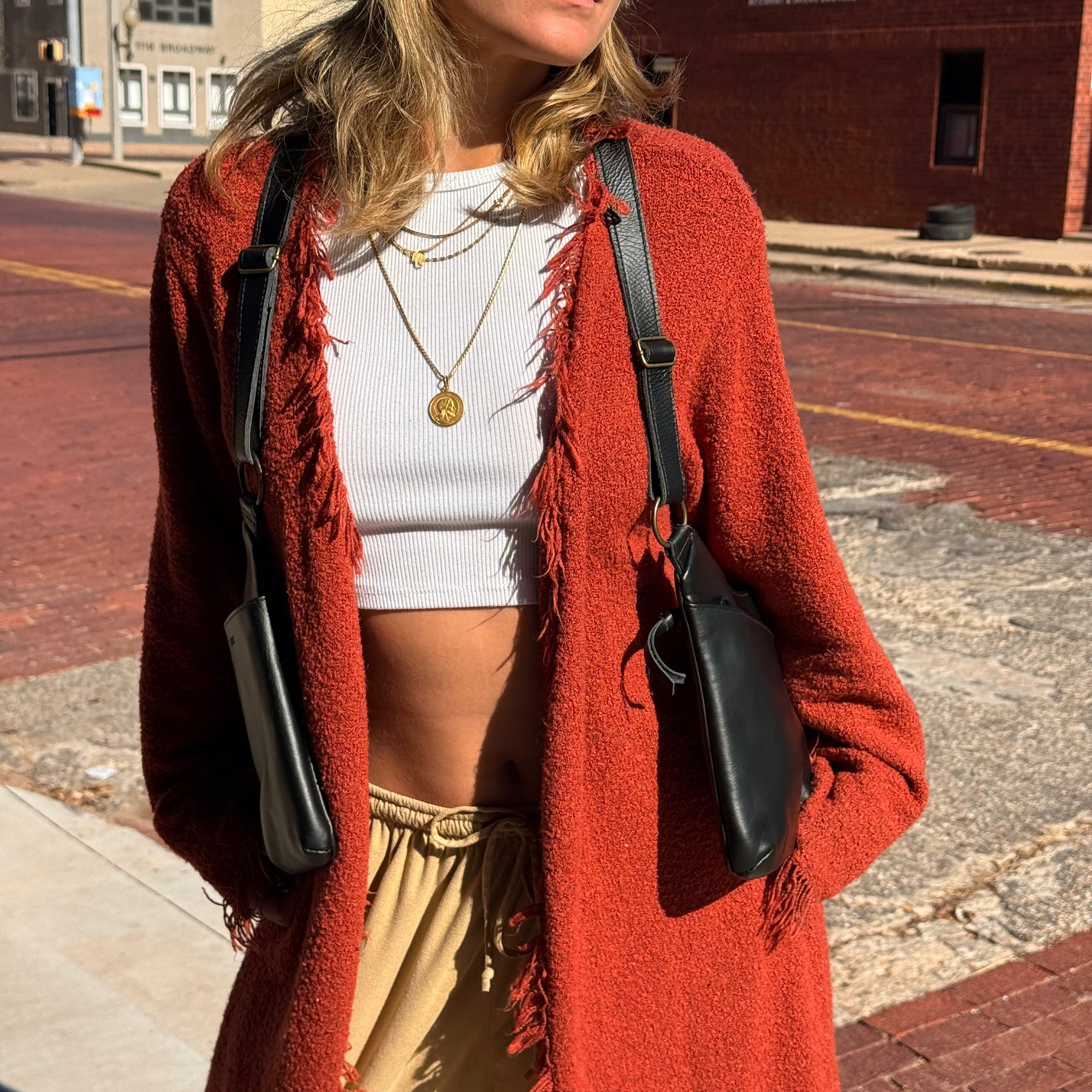Woman in a red cardigan and white top standing on a street corner. Wearing black saddle bags leather handbag