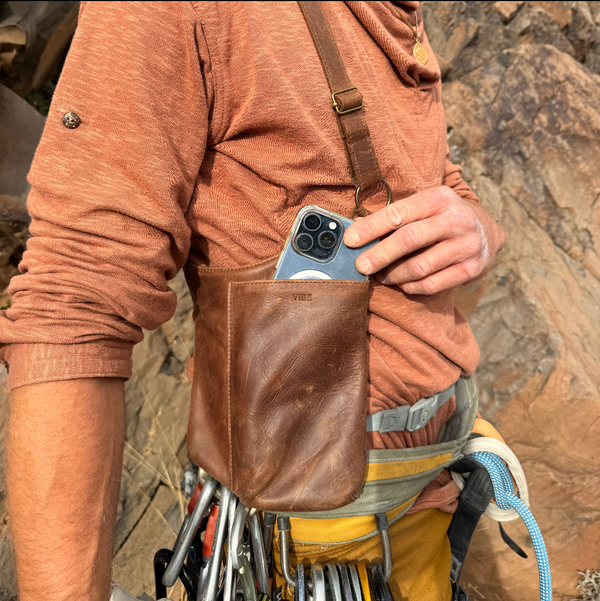 Person in outdoor setting holding a phone in a leather pocket with climbing gear. rock climbers bag, adventure bag, bag for rock climbers