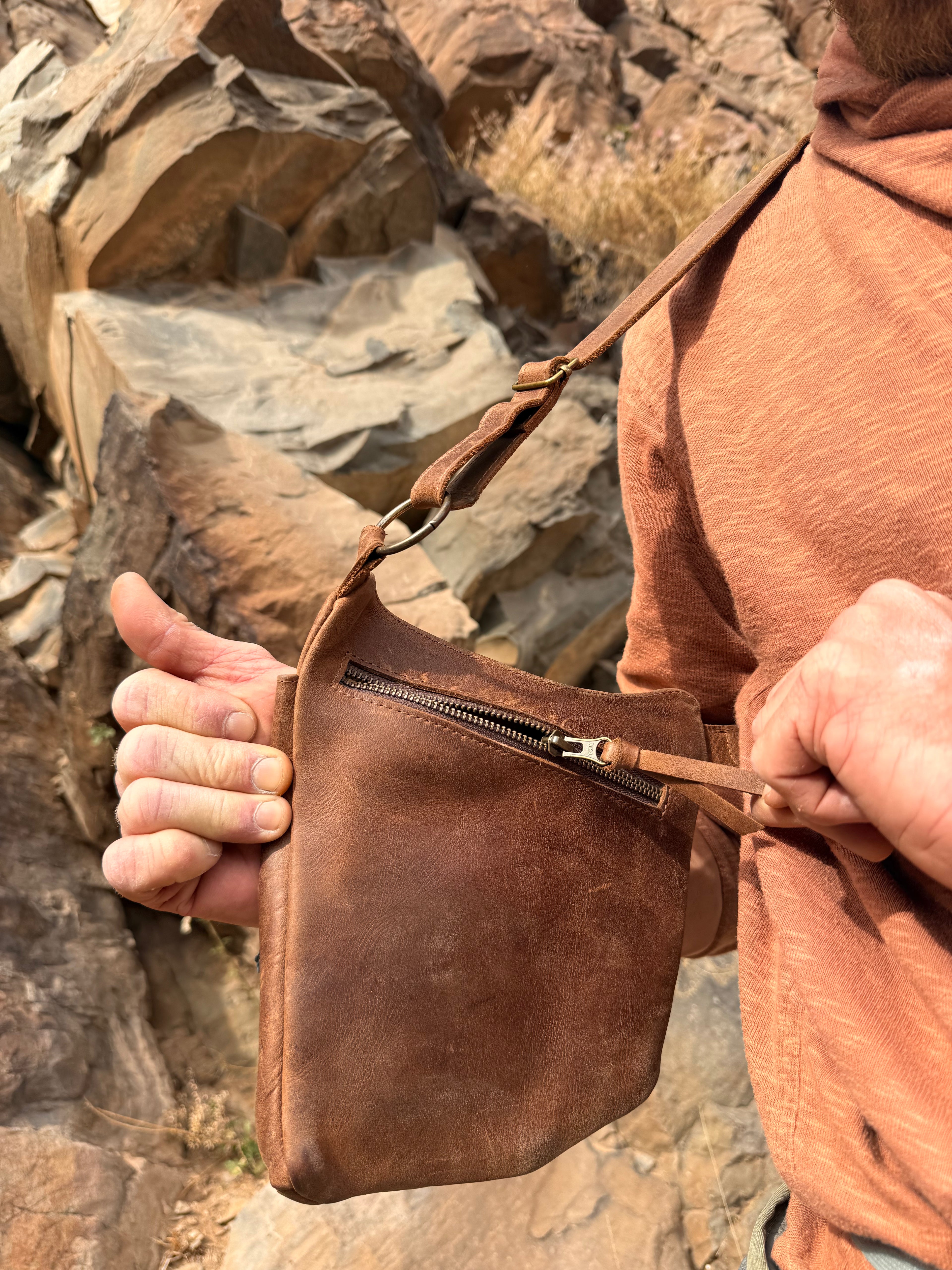 Person holding a brown leather bag against a rocky background. rock climber harness bag, holster bag, leather adventure bag 